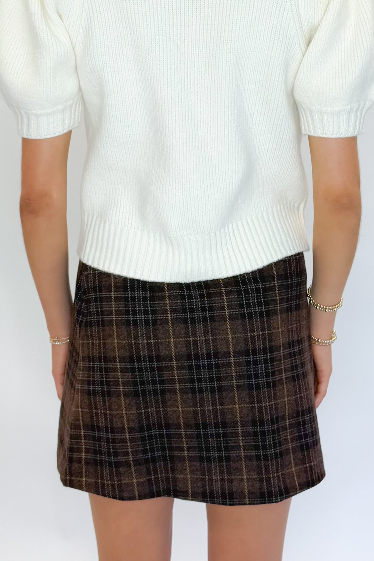Back view of woman wearing Swagger Boutique’s Cream Tie Neck Short Sleeve Sweater by Entro with Brown Plaid Mini Skirt against white studio background.