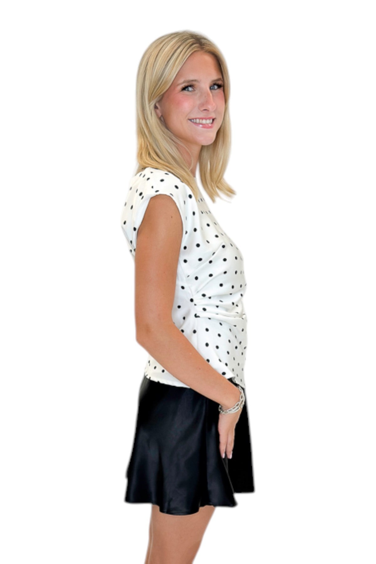 Side view of woman wearing Swagger Boutique’s Cream &amp; Black Polka Dot Boat Neck Tank with Black Satin Mini Skirt against white studio background.