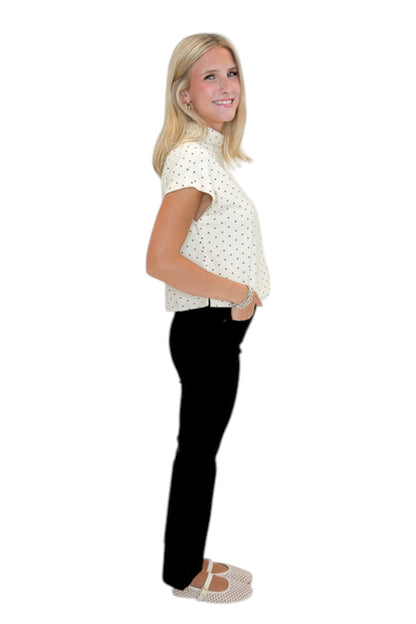 Side view of woman wearing Swagger Boutique’s Cream &amp; Black Polka Dot Mock Neck Top by Entro with jeans against white studio background.
