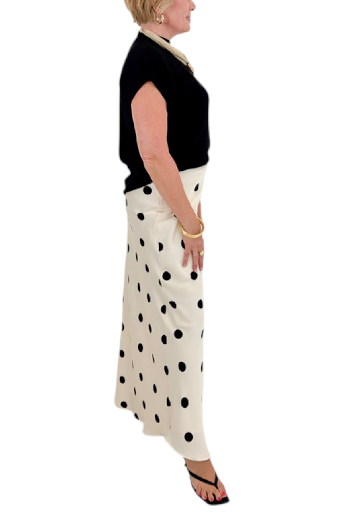 Side detail view of woman wearing Swagger Boutique’s Black Shirred Cap Sleeve Sweater with Cream &amp; Black Polka Dot Satin Skirt against white studio background.