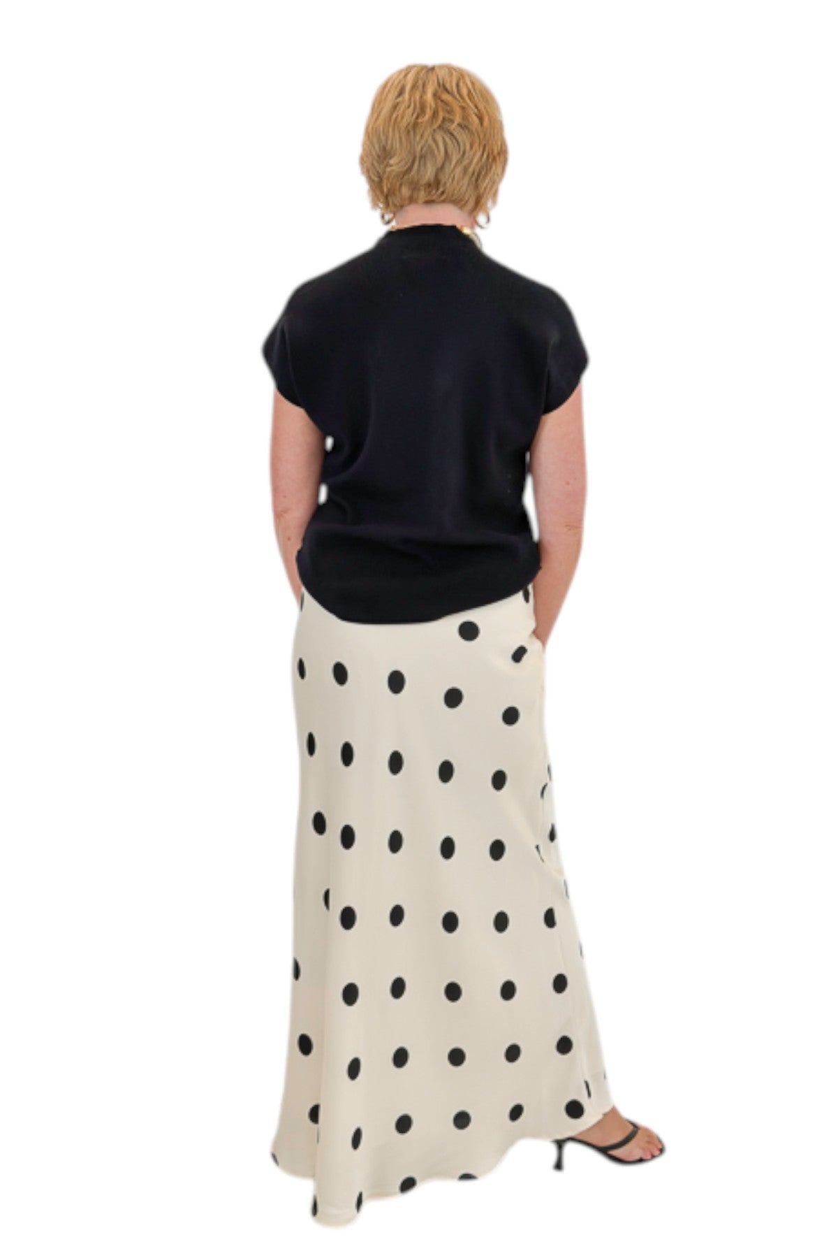 Back view of woman wearing Swagger Boutique’s Black Shirred Cap Sleeve Sweater with Cream &amp; Black Polka Dot Satin Skirt against white studio background.