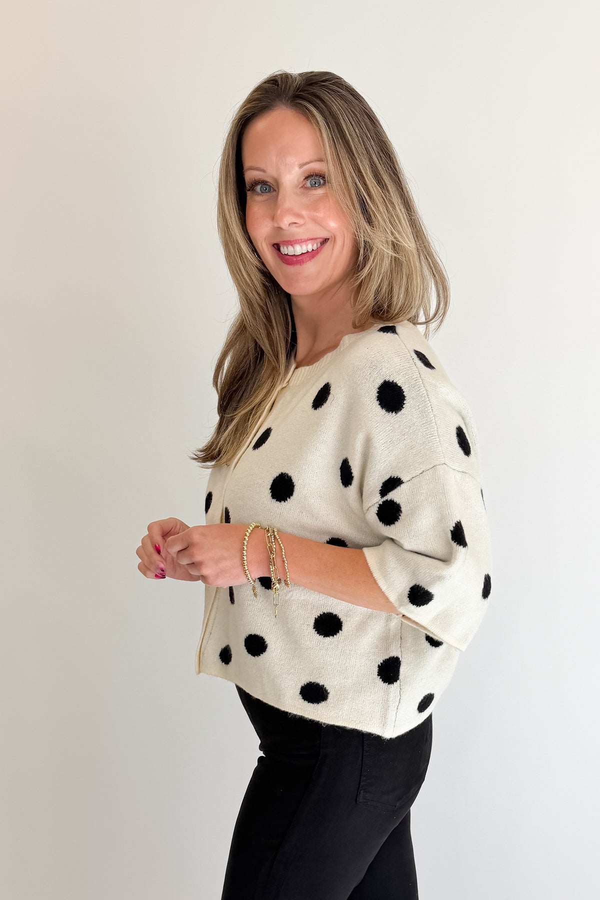 Side view of woman wearing Swagger Boutique’s Cream &amp; Black Polka Dot Short Sleeve Cardigan with jeans against white studio background.