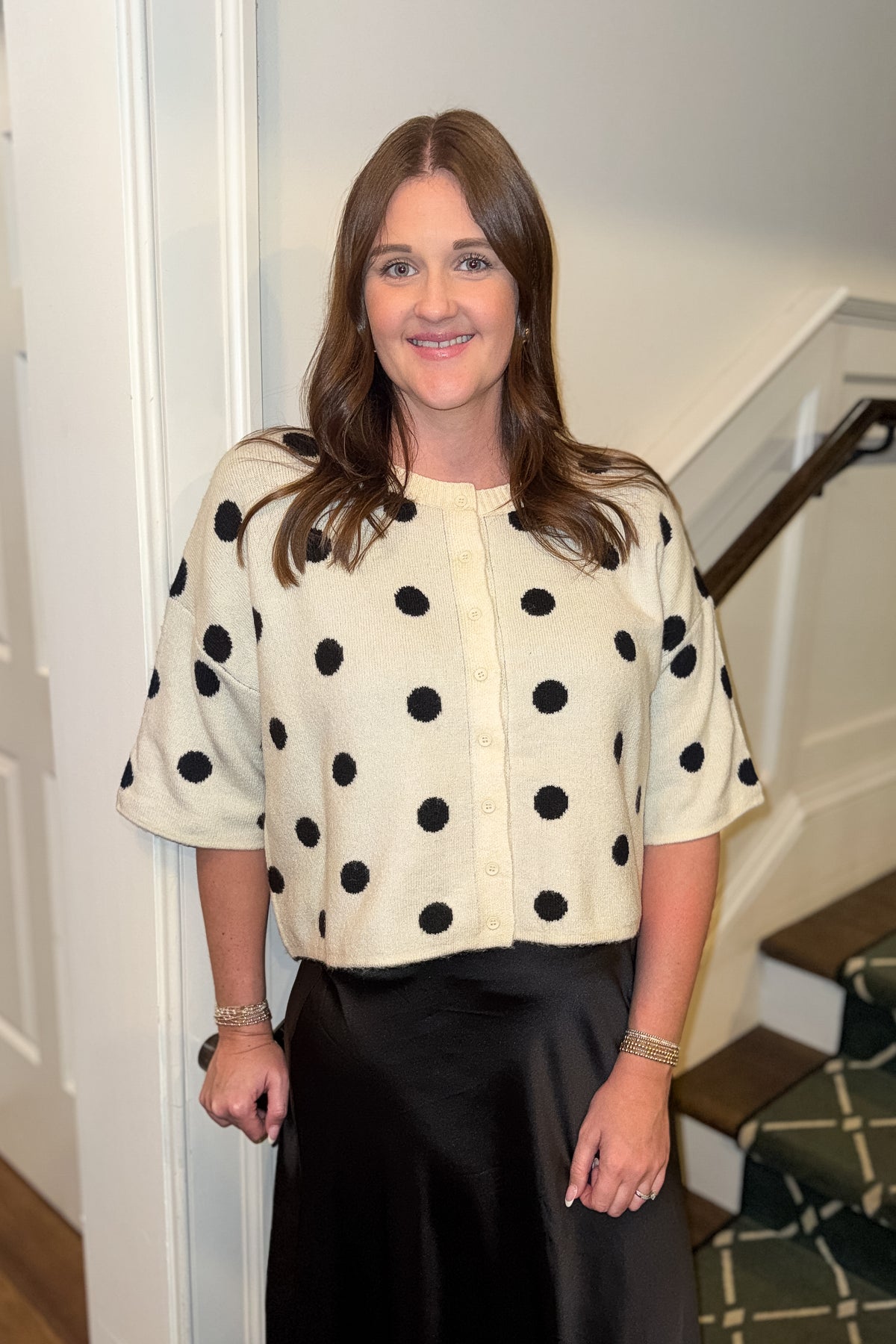 Front view of woman wearing Swagger Boutique’s Cream &amp; Black Polka Dot Short Sleeve Cardigan with skirt standing by a staircase.
