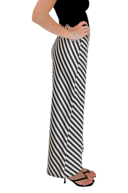 Side view of woman wearing Swagger Boutique’s Cream &amp; Black Striped Satin Midi Skirt with black top against white studio background.