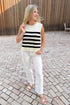 Front view of model wearing Cream & Black Striped Sleeveless Sweater by Entro and jeans standing outside on a brick path.