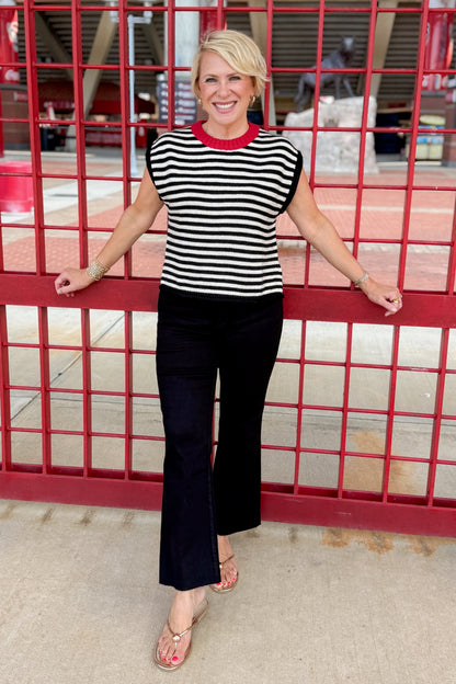 Front view of woman wearing Swagger Boutique’s Cream &amp; Black Striped Sleeveless Sweater with jeans standing at a stadium gate. 