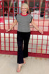 Front view of woman wearing Swagger Boutique’s Cream & Black Striped Sleeveless Sweater with jeans standing at a stadium gate. 