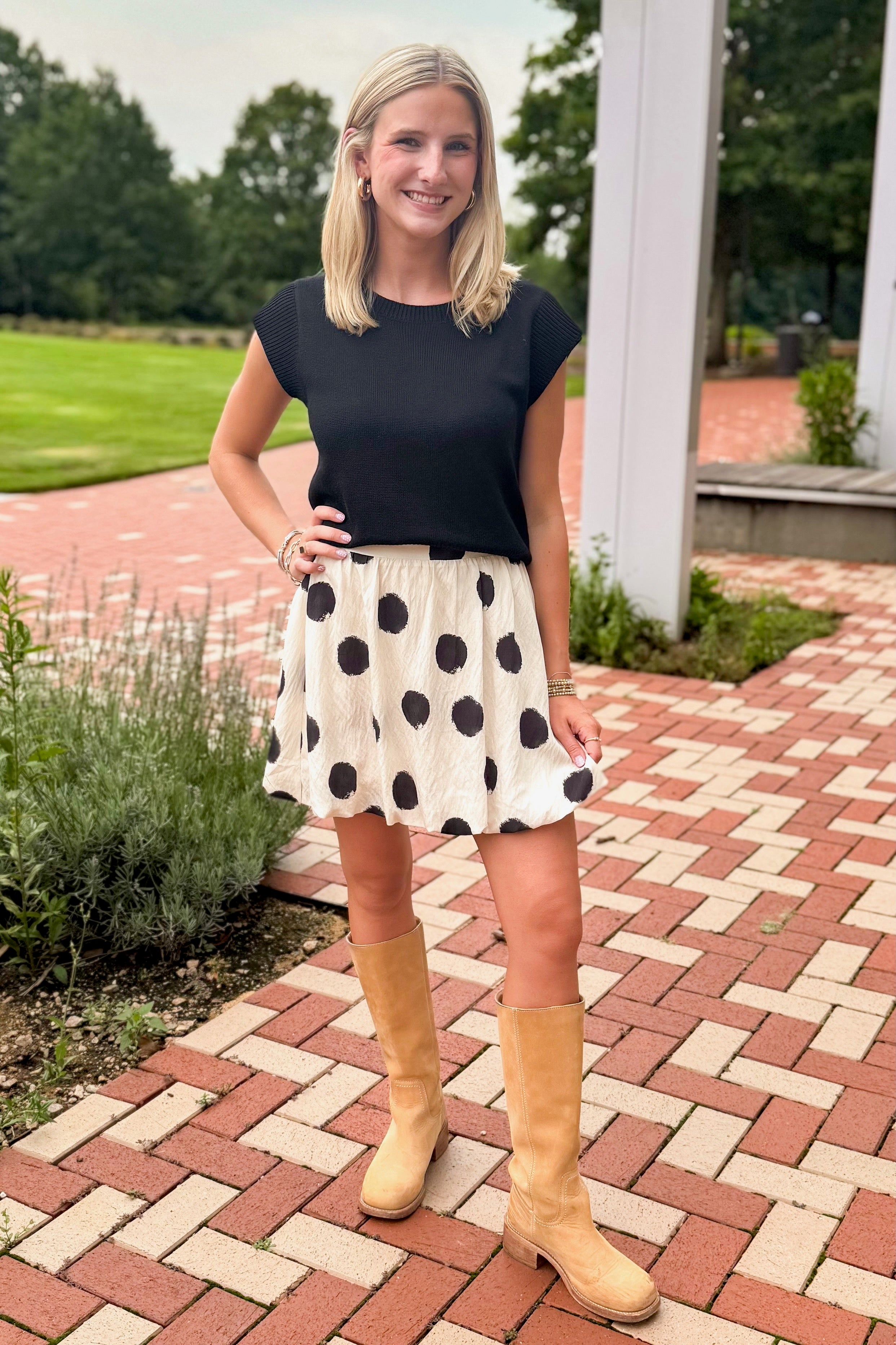 Front view of woman wearing Swagger Boutique’s Black Cap Sleeve Sweater with polka dot skirt standing on a brick path outside.