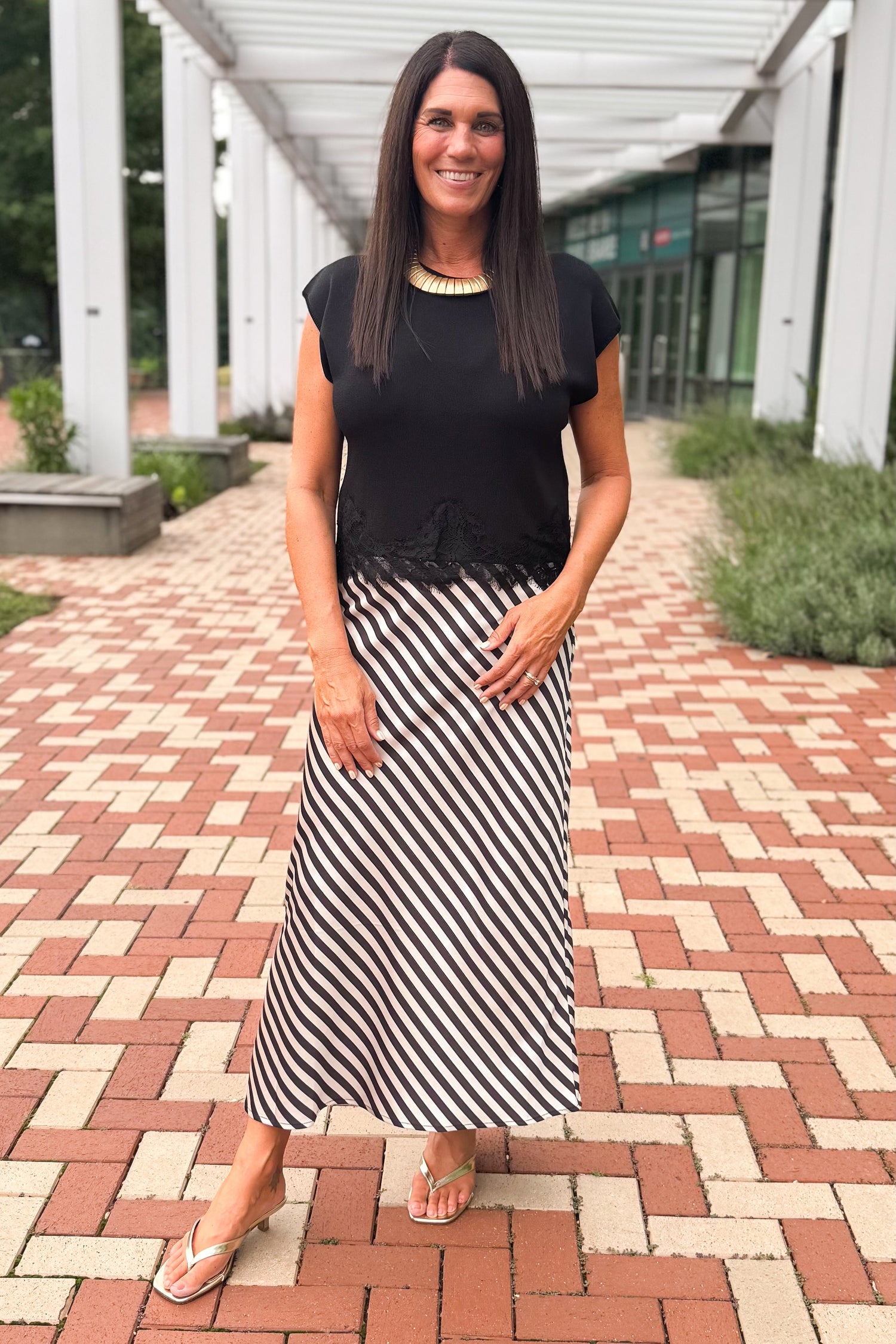 Front view of woman wearing Swagger Boutique’s Black Short Sleeve Lace Trim Top with stripe skirt at a brick pathway outside.