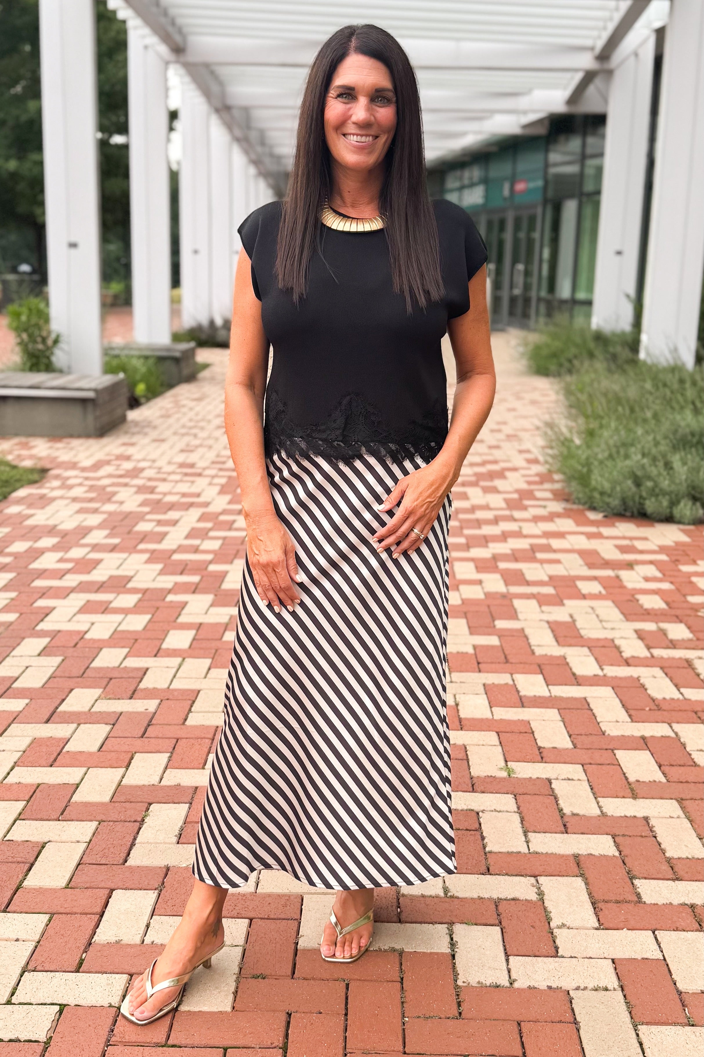 Front view of woman wearing Swagger Boutique’s Black Short Sleeve Lace Trim Top with stripe skirt at a brick pathway outside.