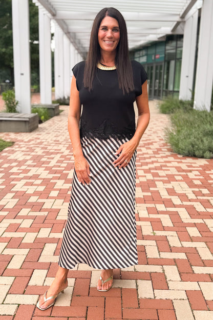Front view of woman wearing Swagger Boutique’s Black Short Sleeve Lace Trim Top with stripe skirt at a brick pathway outside.
