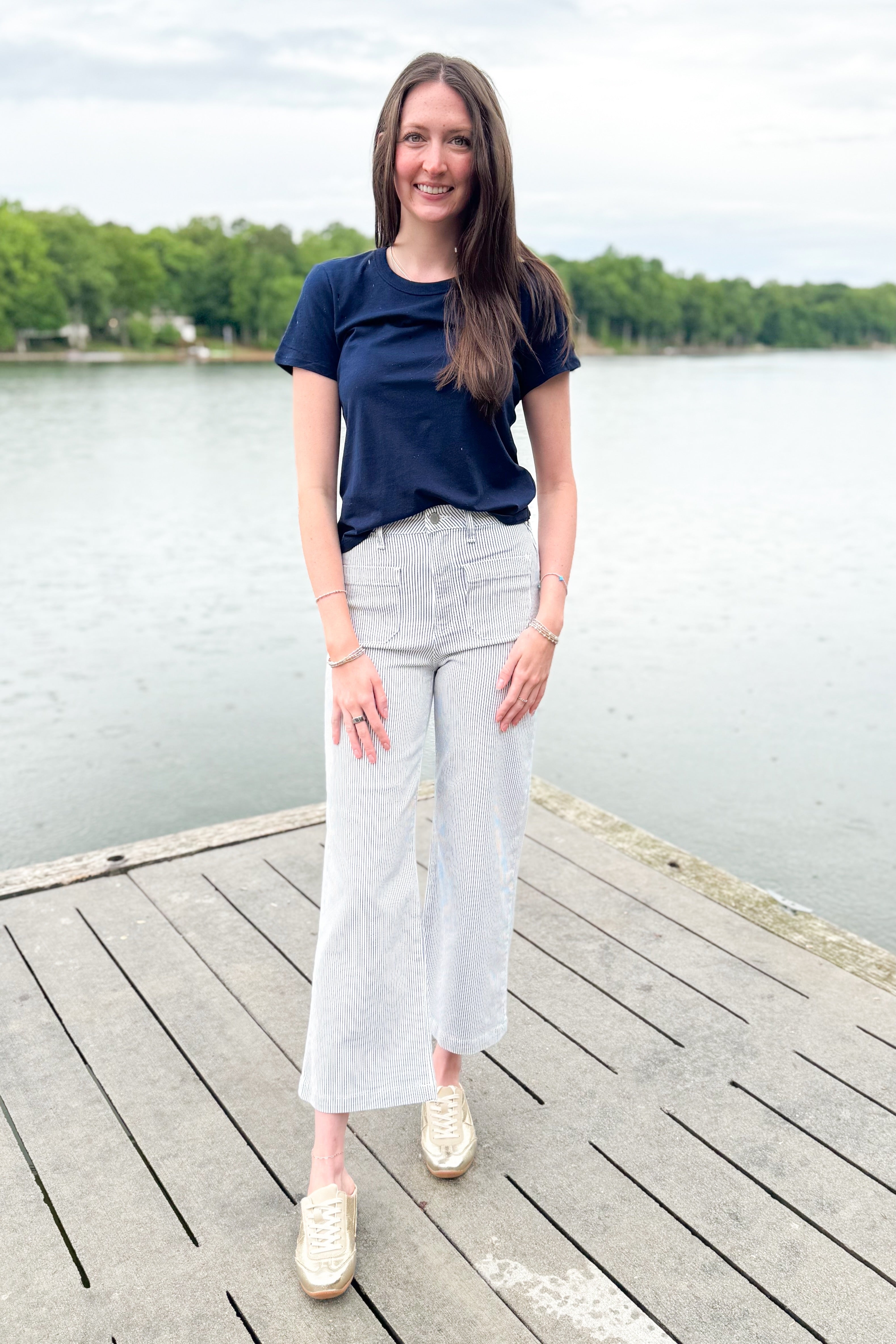 Front view of woman wearing Swagger Boutique’s Cream &amp; Blue Striped Jeans With Patch Pocket by Just Black Denim with blue Z Supply top outside at a lake.