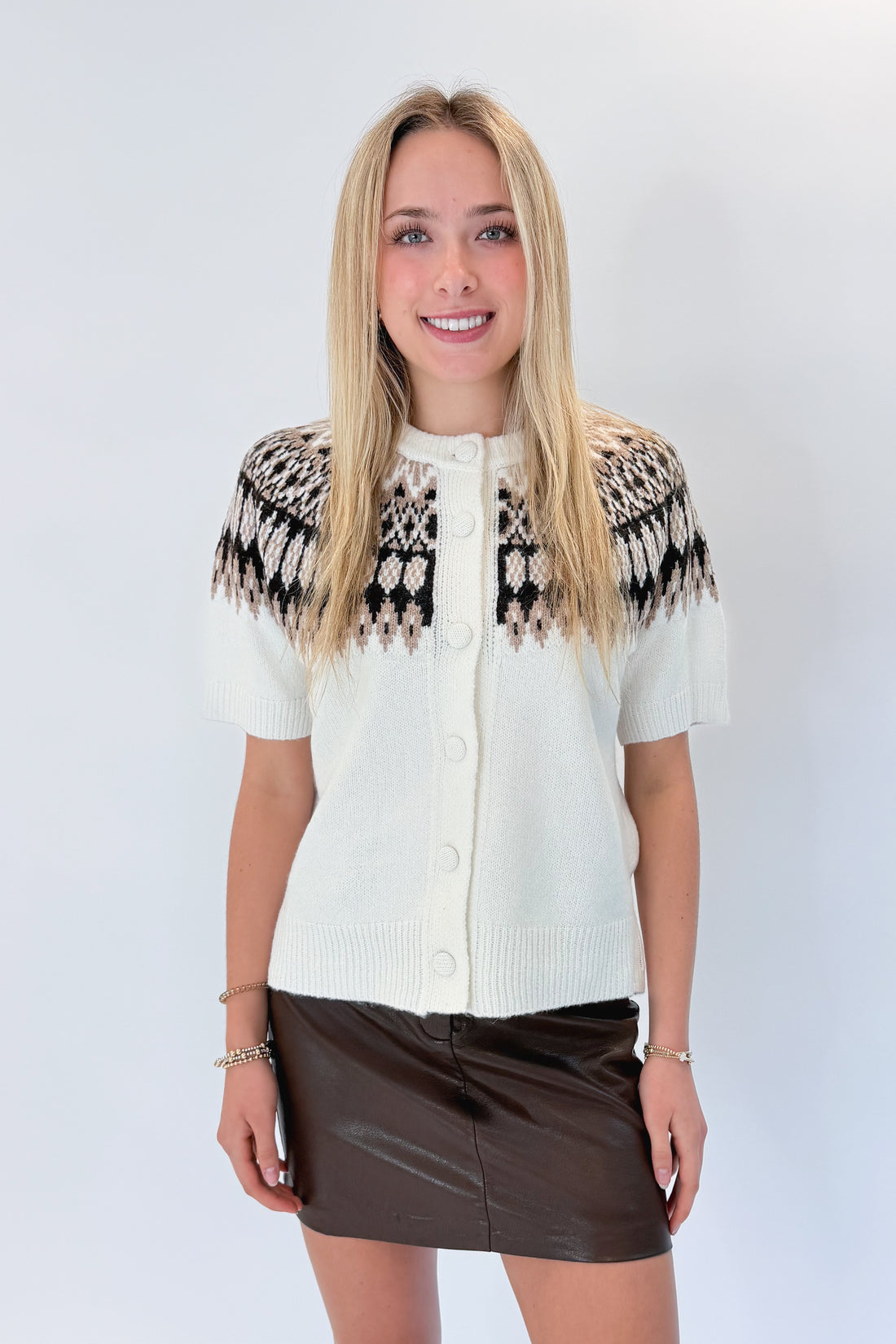 Front view of woman wearing Swagger Boutique’s Cream &amp; Brown Fair Isle Button Up Short Sleeve Sweater with brown faux leather mini skirt against white studio background. 