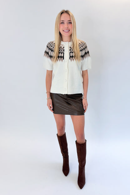 Front view of woman wearing Swagger Boutique’s Cream &amp; Brown Fair Isle Button Up Short Sleeve Sweater with brown faux leather mini skirt against white studio background. 