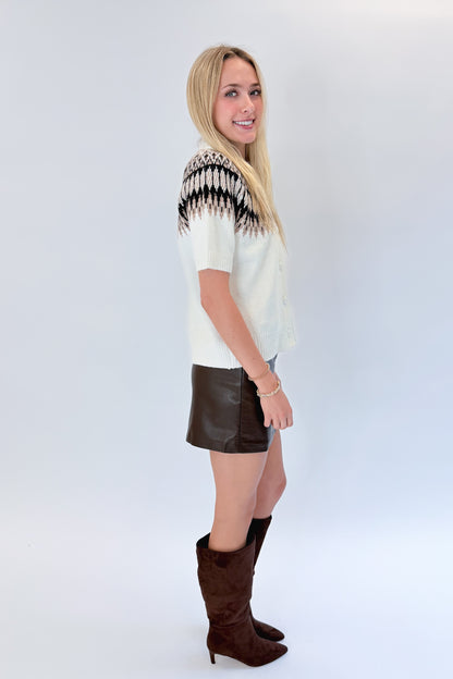 Side view of woman wearing Swagger Boutique’s Cream &amp; Brown Fair Isle Button Up Short Sleeve Sweater with brown faux leather mini skirt against white studio background. 