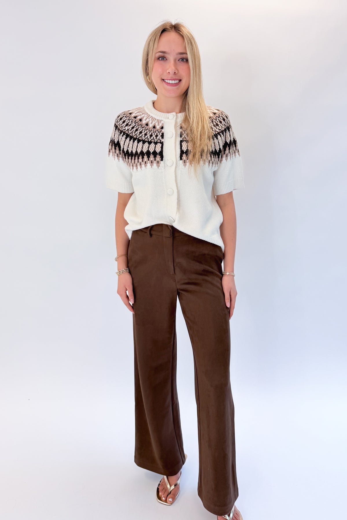 Front view of woman wearing Swagger Boutique’s Cream &amp; Brown Fair Isle Button Up Short Sleeve Sweater with Chocolate Brown Suede Pants against white studio background. 