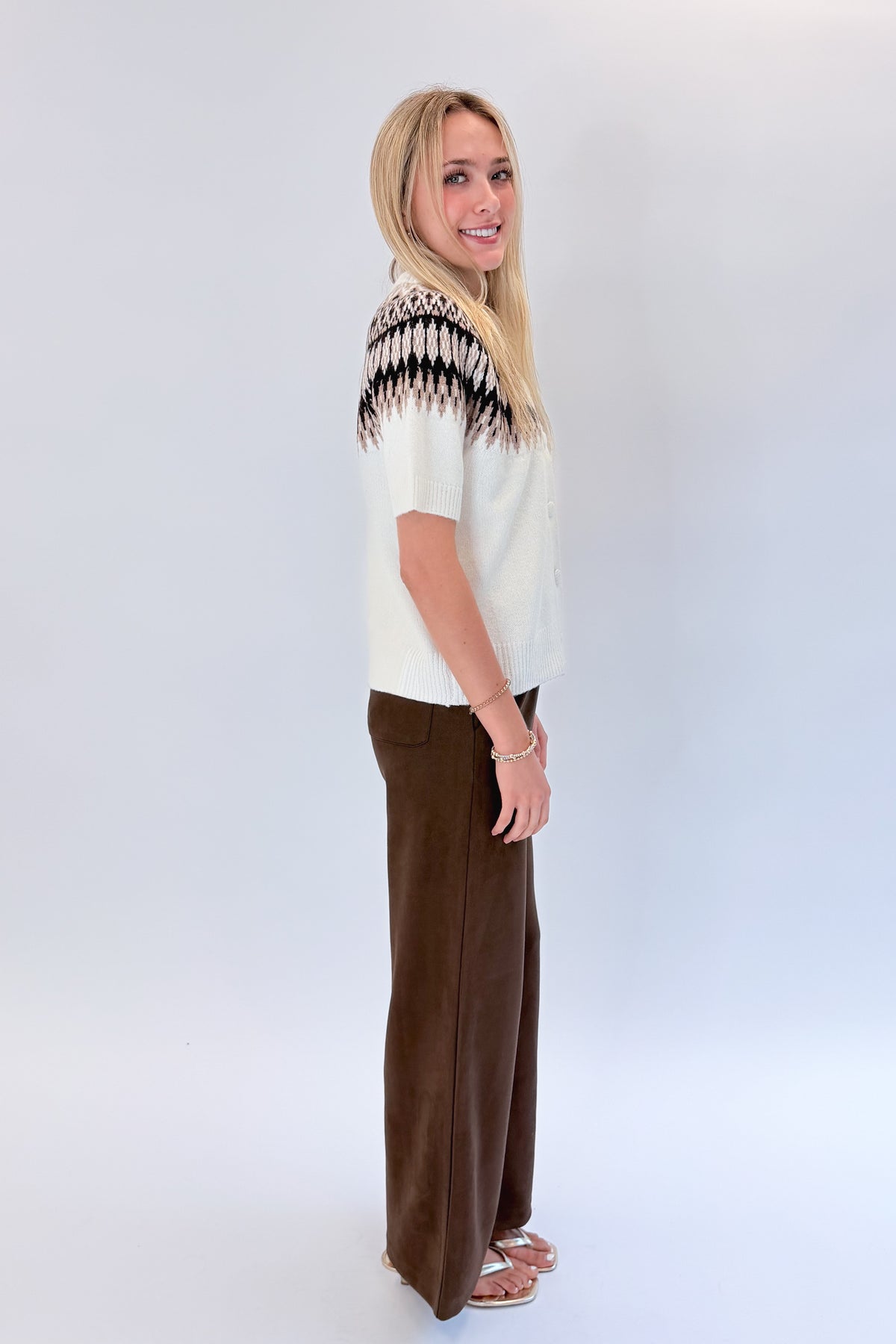 Side view of woman wearing Swagger Boutique’s Cream &amp; Brown Fair Isle Button Up Short Sleeve Sweater with Chocolate Brown Suede Pants against white studio background. 
