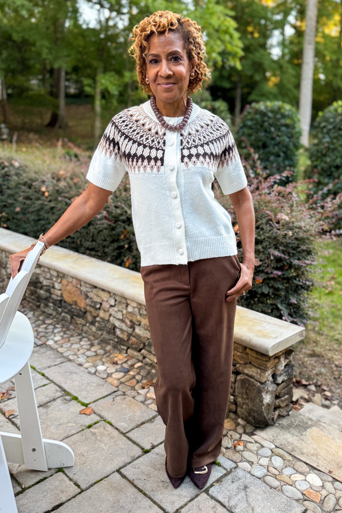 Front view of woman wearing Swagger Boutique’s Cream &amp; Brown Fair Isle Button Up Short Sleeve Sweater with Chocolate Brown Suede Pants standing outside at a party.