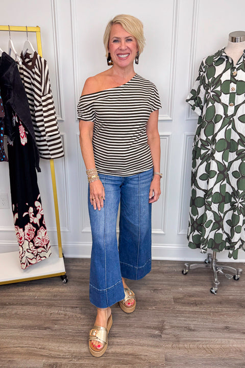 Front view of woman wearing Swagger Boutique’s Cream &amp; Brown Striped Asymmetrical Top with jeans with clothing hanging in the background.
