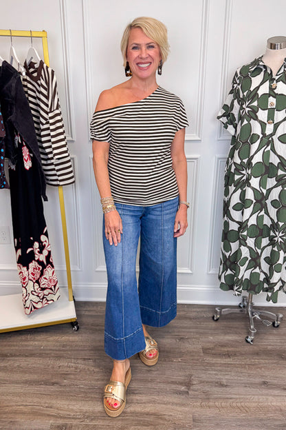 Front view of woman wearing Swagger Boutique’s Cream &amp; Brown Striped Asymmetrical Top with jeans with clothing hanging in the background.