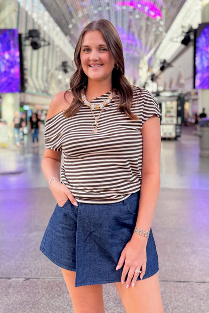A woman posing in a modern indoor shopping center or airport terminal, wearing a brown and white horizontally striped off-the-shoulder top and a dark blue denim wrap skirt.