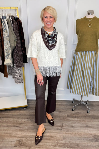 Front view of woman wearing Swagger Boutique’s Cream Fringe Short Sleeve Sweater with jeans with clothing hanging in the background.