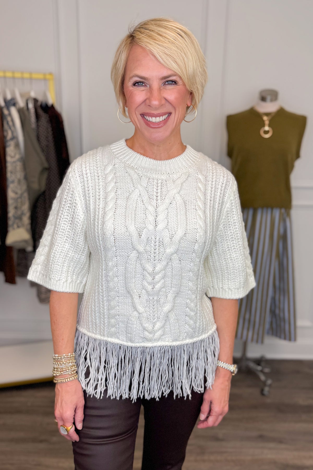 Front view of woman wearing Swagger Boutique’s Cream Fringe Short Sleeve Sweater with jeans with clothing hanging in the background.