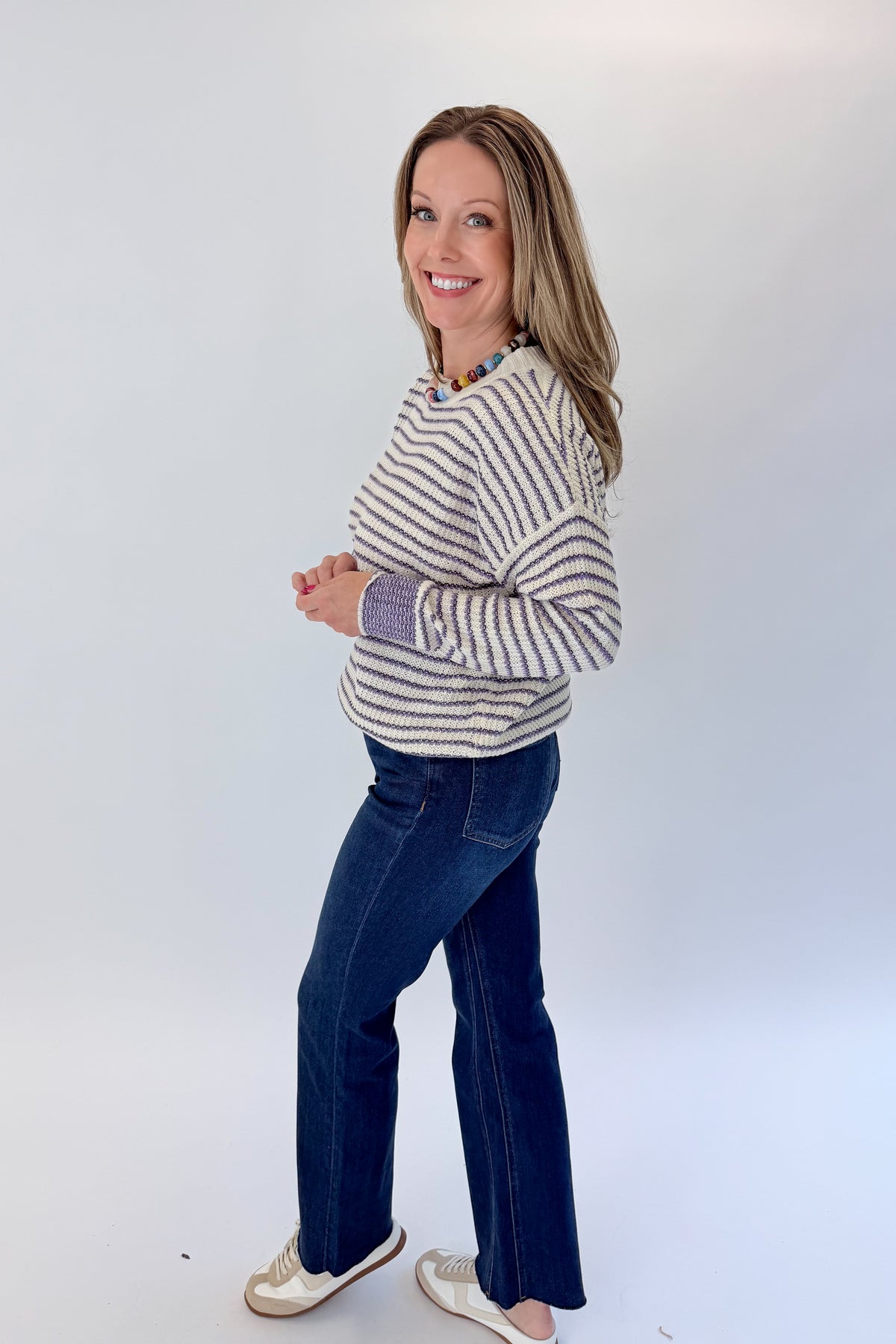 Side view of woman wearing Swagger Boutique’s Cream &amp; Purple Striped Sweater with jeans against white studio background.