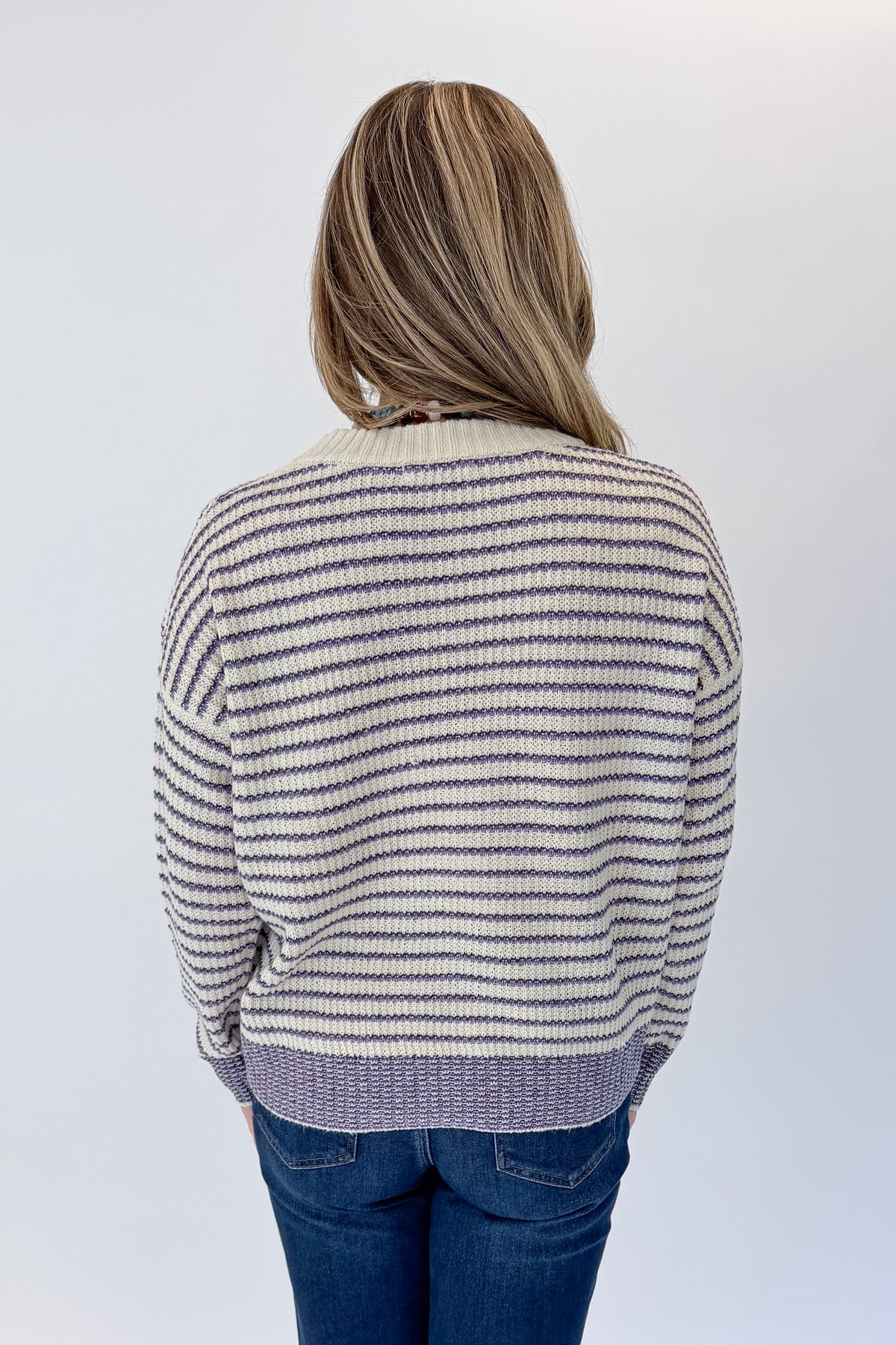 Back view of woman wearing Swagger Boutique’s Cream &amp; Purple Striped Sweater with jeans against white studio background.