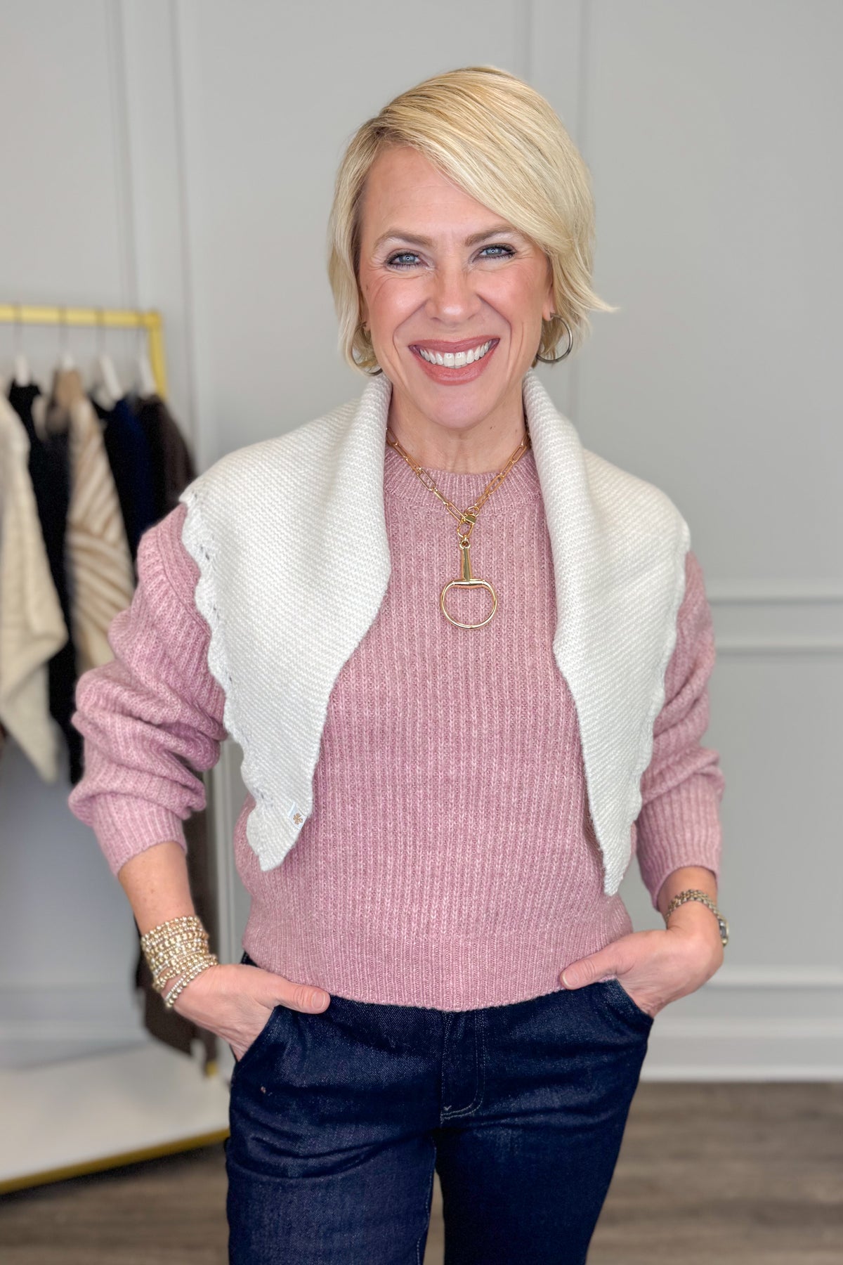 A smiling blonde woman wearing a pink ribbed knit sweater, a white knit shrug vest with ruffled edges, and dark blue jeans, standing in a boutique setting.
