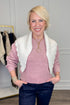 A smiling blonde woman wearing a pink ribbed knit sweater, a white knit shrug vest with ruffled edges, and dark blue jeans, standing in a boutique setting.