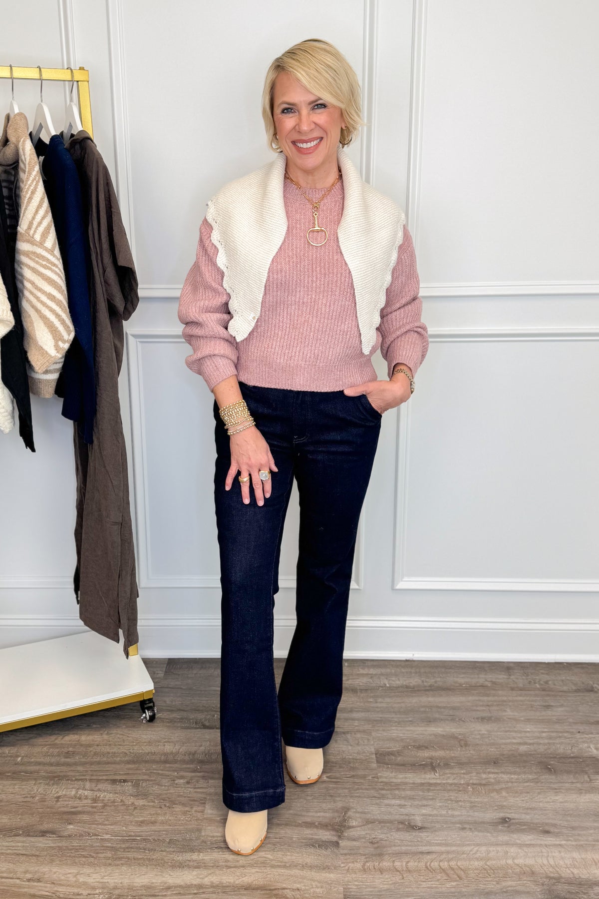 A woman stands indoors modeling a pink knit sweater vest layered over a cream faux-fur vest, paired with dark blue flared jeans and beige flats. She is posed in front of a white paneled wall with a clothing rack visible on the left.
