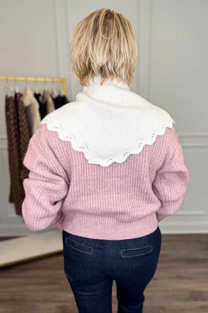 A woman viewed from behind wearing a pink ribbed knit sweater with a large white scalloped knit scarf paired with dark blue jeans in an indoor boutique setting.