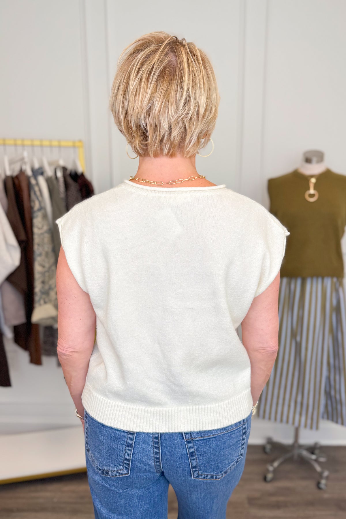 Back view of woman wearing Swagger Boutique’s Cream Sleeveless Button Up Sweater with Maya Denim Jeans by Kut from the Kloth with clothing hanging in the background.