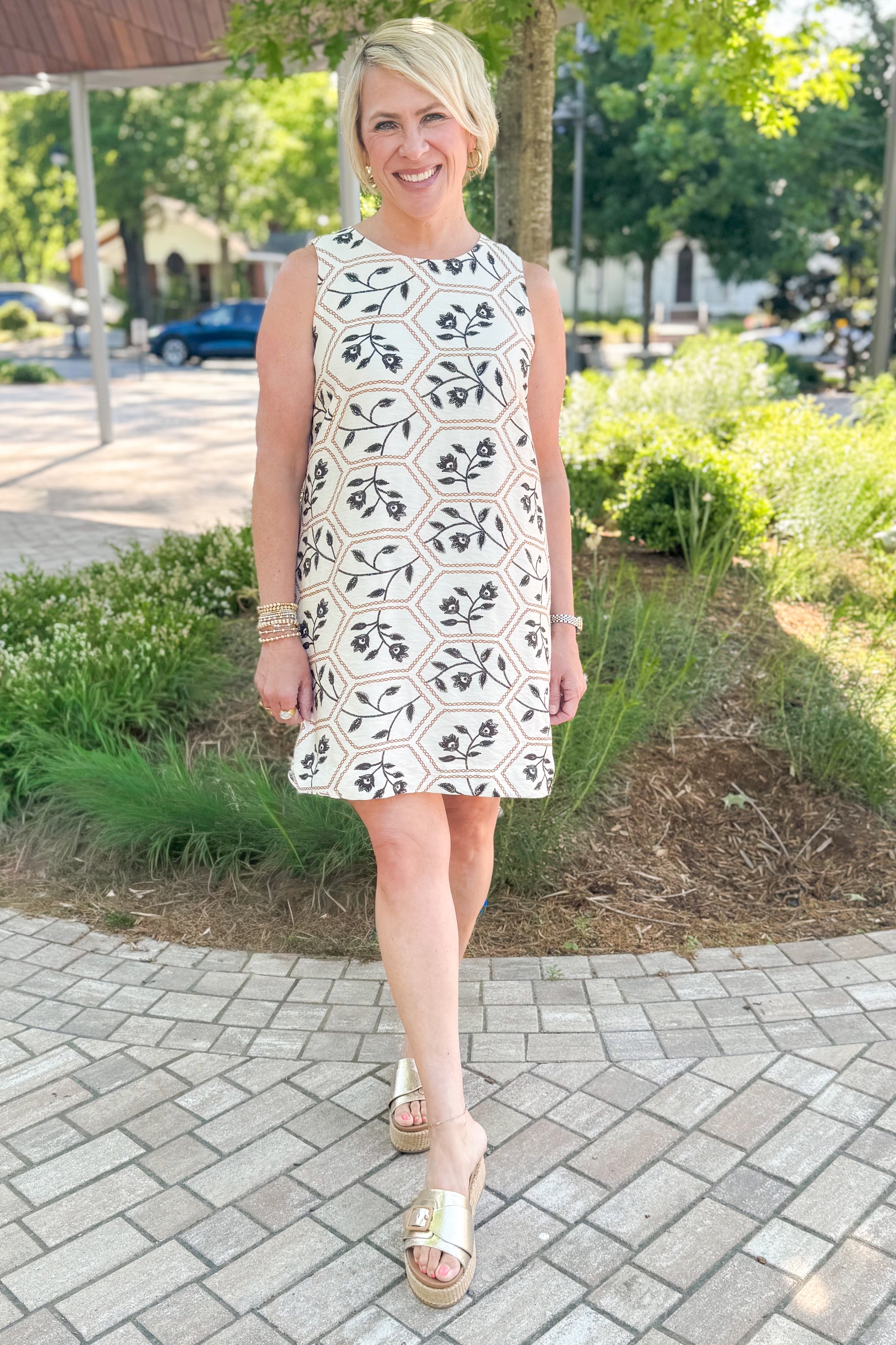 Front view of woman wearing Swagger Boutique’s Cream, Taupe &amp; Black Floral Printed Shift Dress with sandals outside at a park.