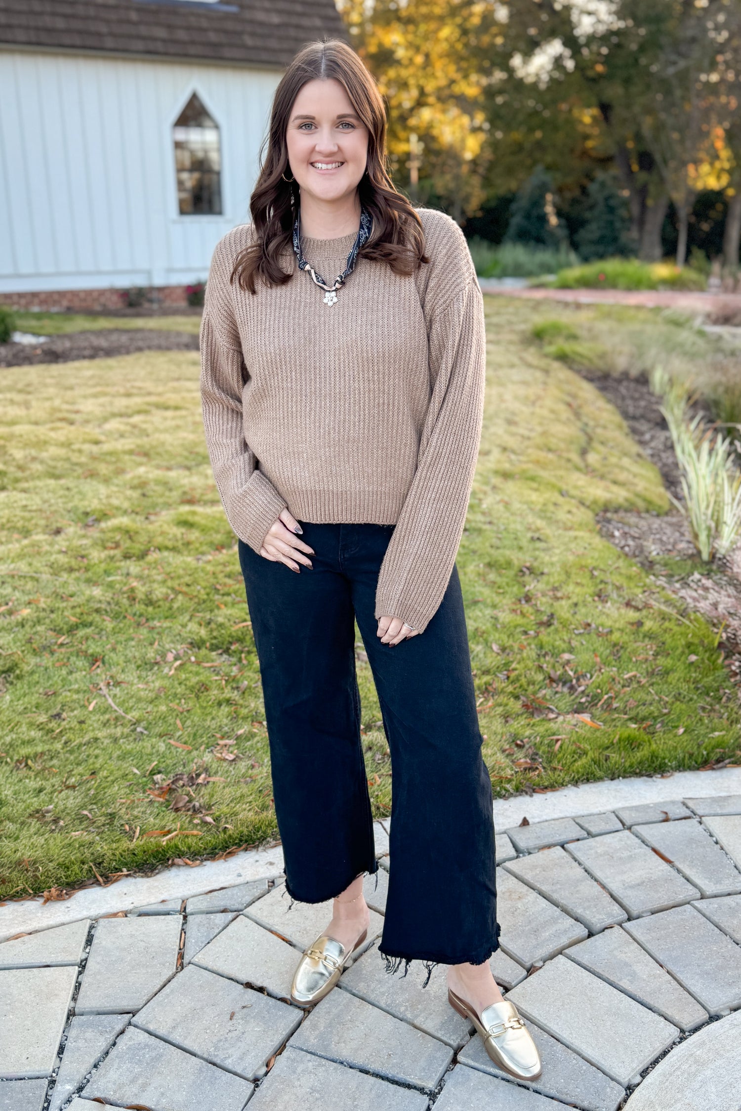 Woman in a beige sweater and black pants standing on a paved walkway with grass and trees in the background.
