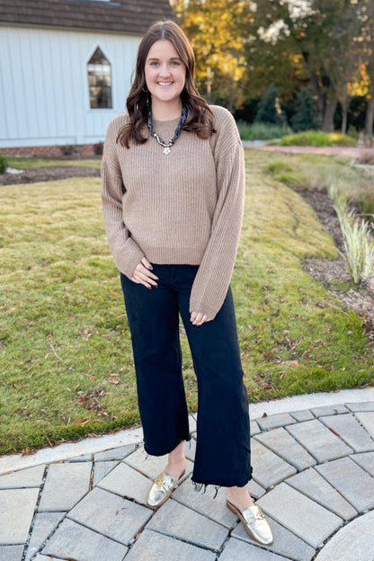 Woman in a beige sweater and black pants standing on a paved walkway with grass and trees in the background.