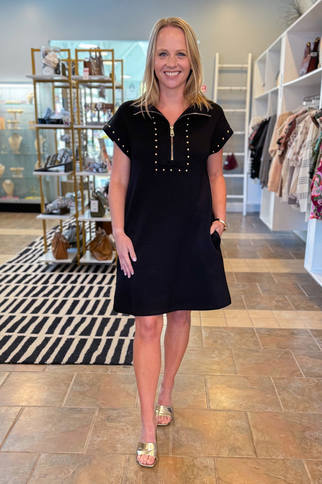 Woman in a black dress standing in a store with clothing racks and displays in the background.
