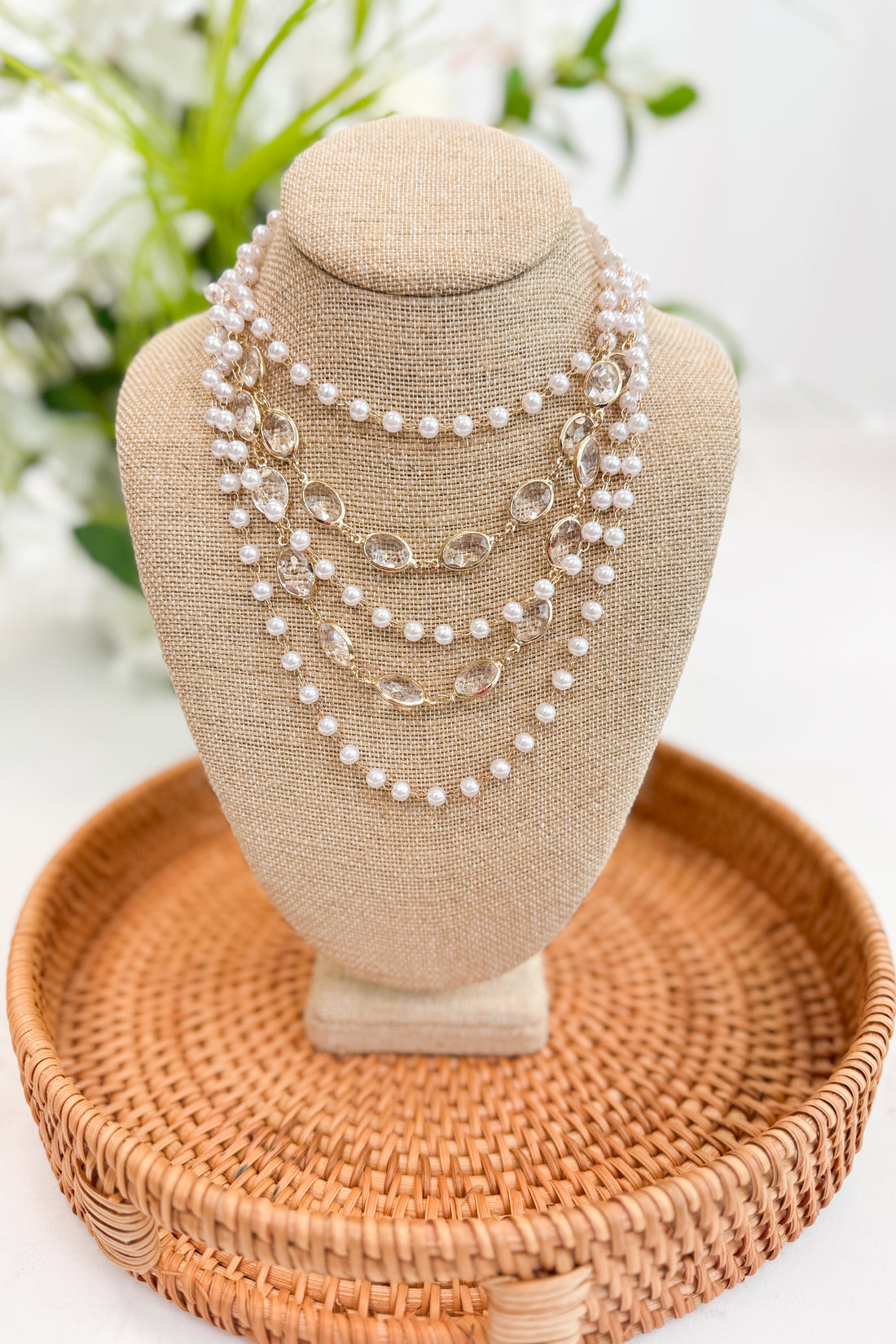 Set of pearl necklaces on a beige bust with a woven base, against a blurred floral background.