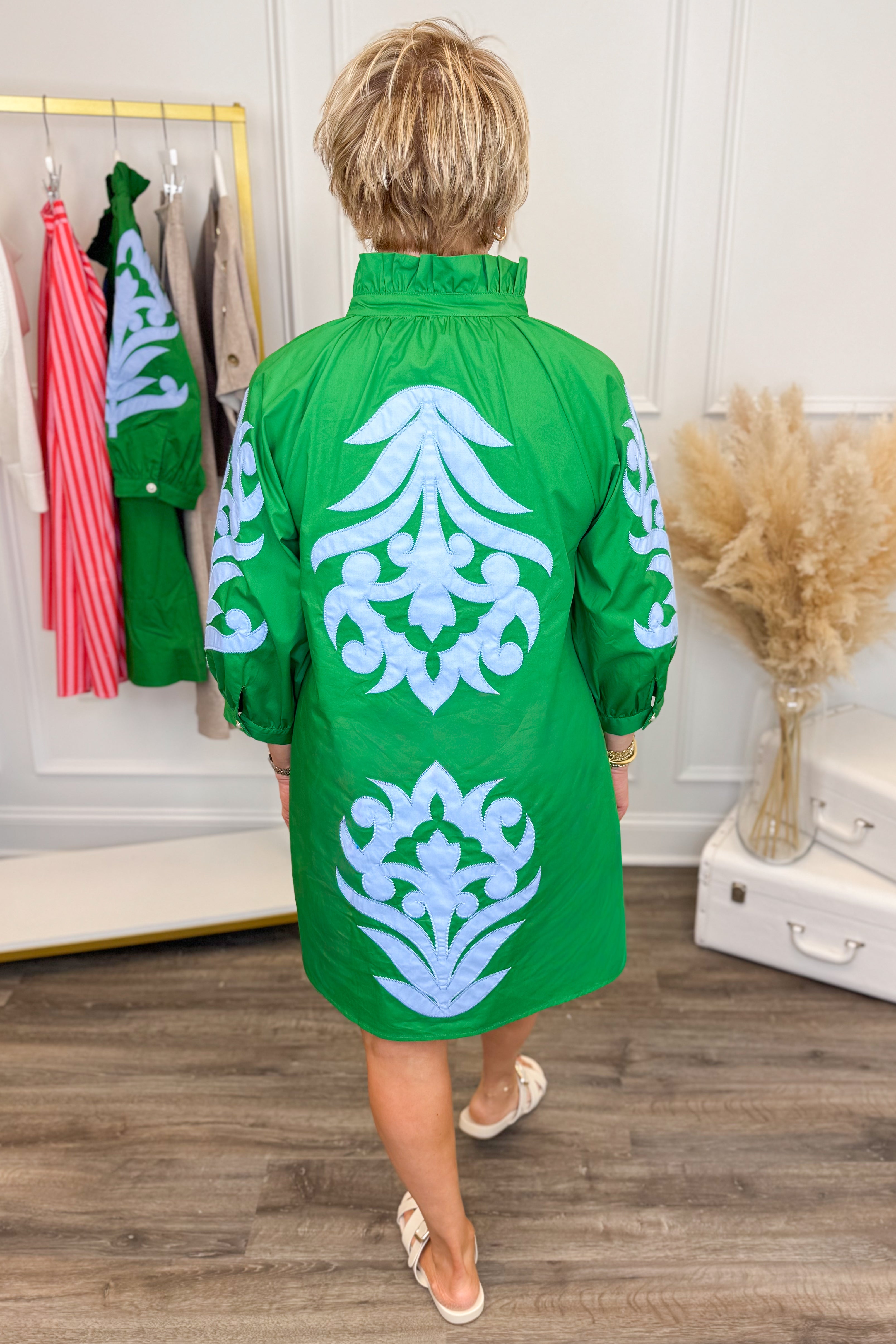 Person wearing a green dress with white floral patterns in a room with clothes and decor.