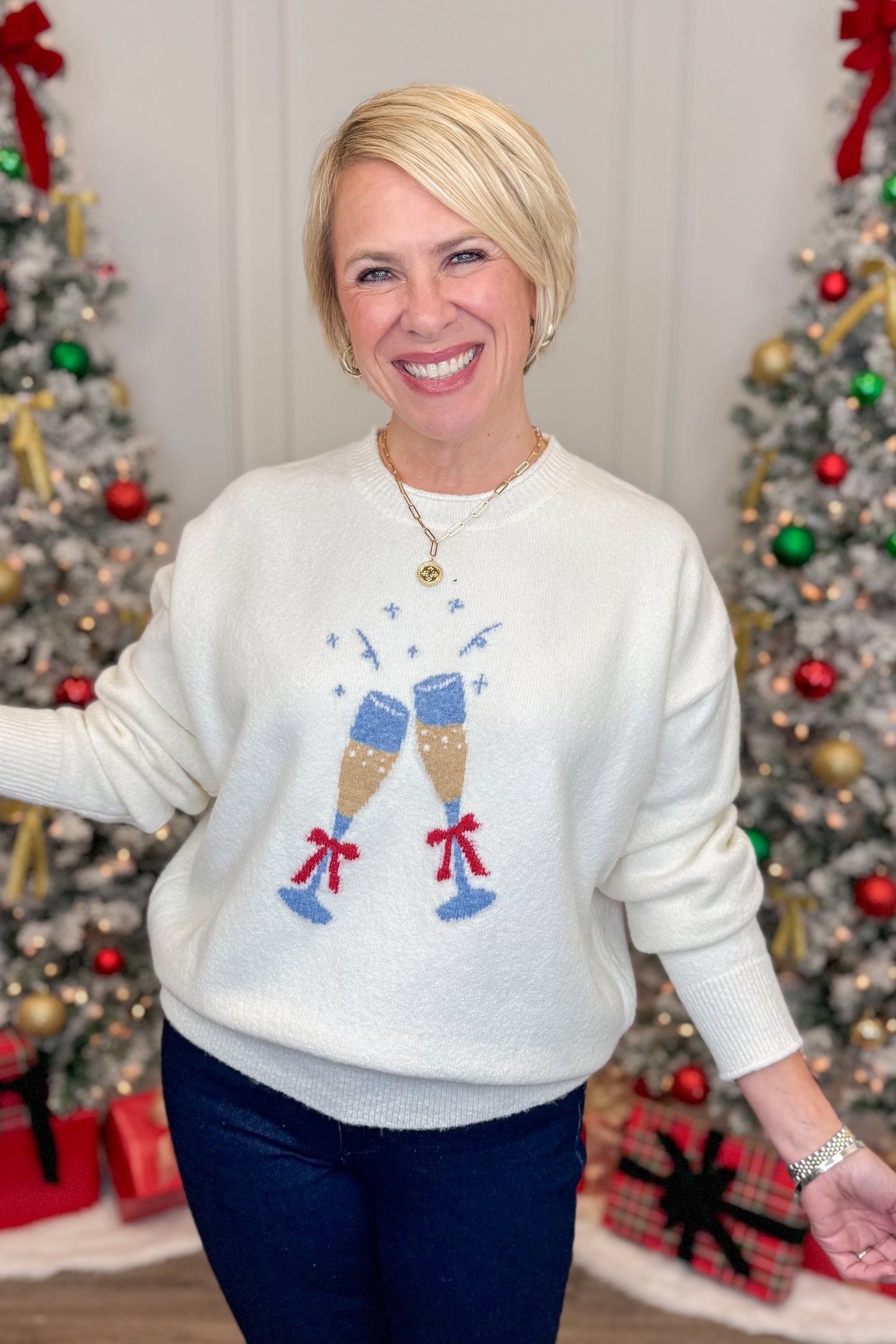 Woman wearing a white sweater with festive design in front of Christmas trees.