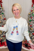 Woman wearing a white sweater with festive design in front of Christmas trees.