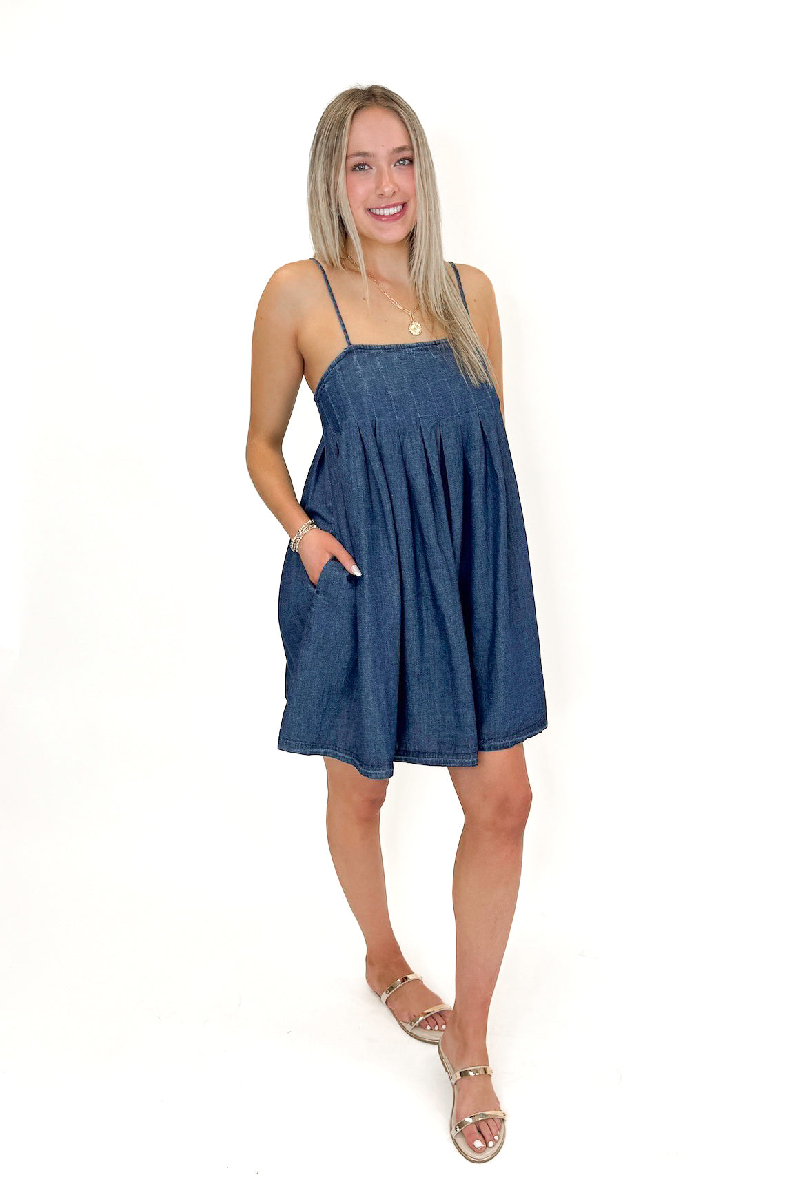 Side view of woman wearing Denim Pleat Detail Tank Dress with sandals against white studio background.
