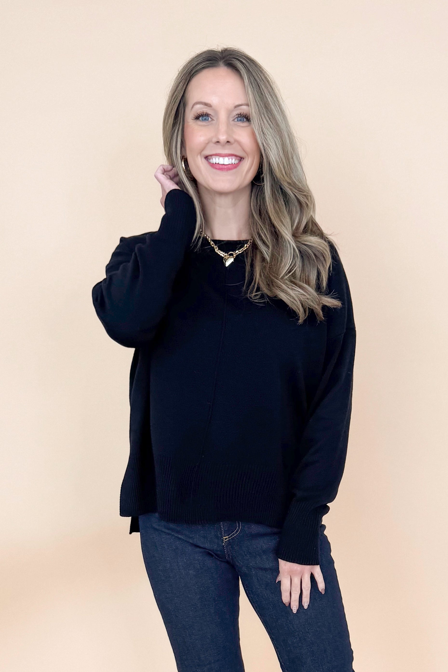 Front view of woman wearing black long sleeve sweater with jeans against tan studio background.