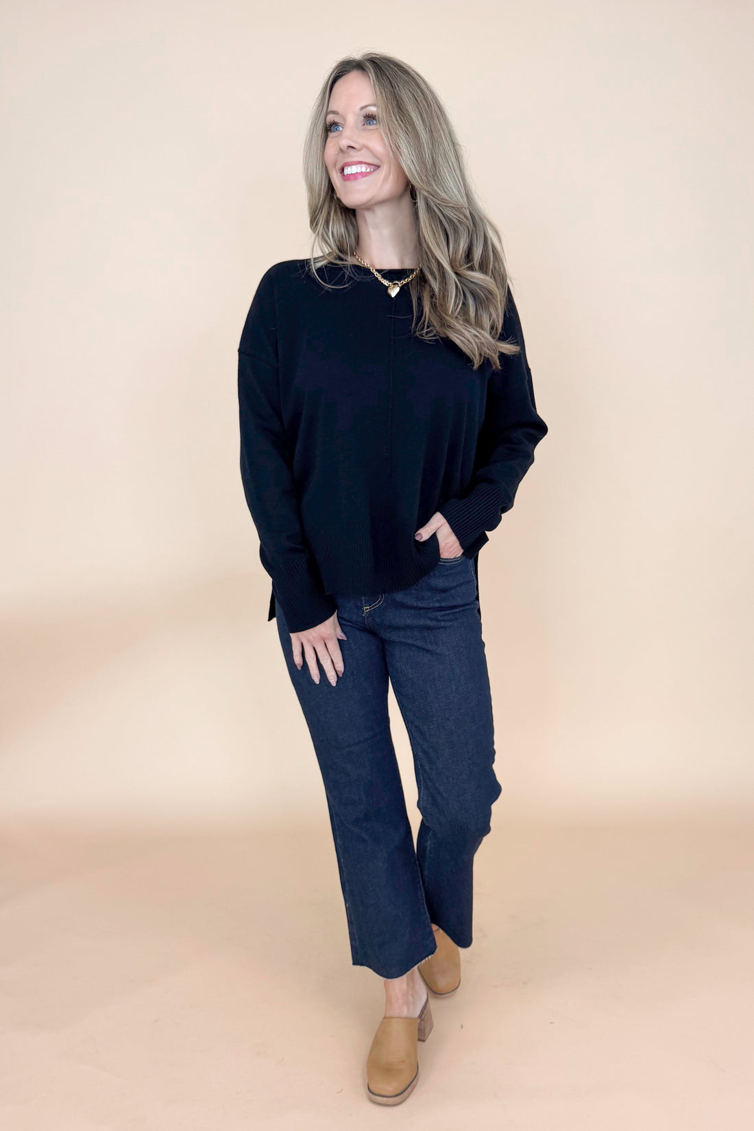 Front view of woman wearing black long sleeve sweater with jeans against tan studio background.