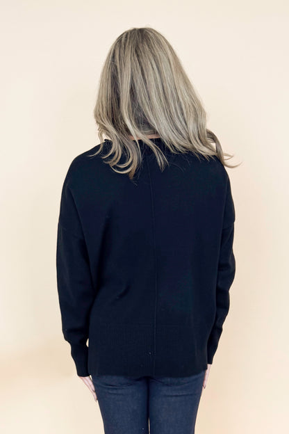 Back view of woman wearing black long sleeve sweater with jeans against tan studio background.