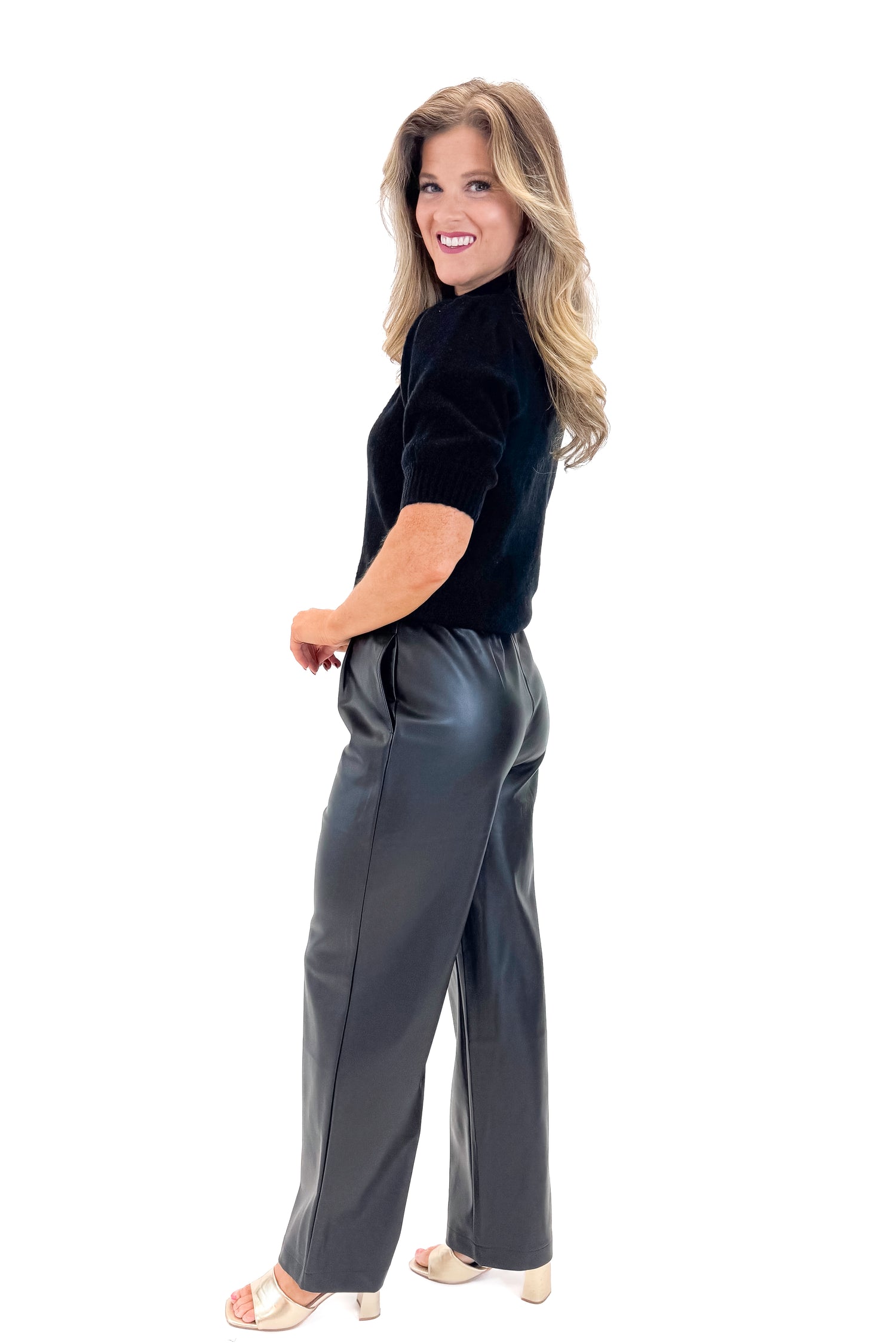 Side view of woman wearing Black Faux Leather Pants with black sweater against white studio background.