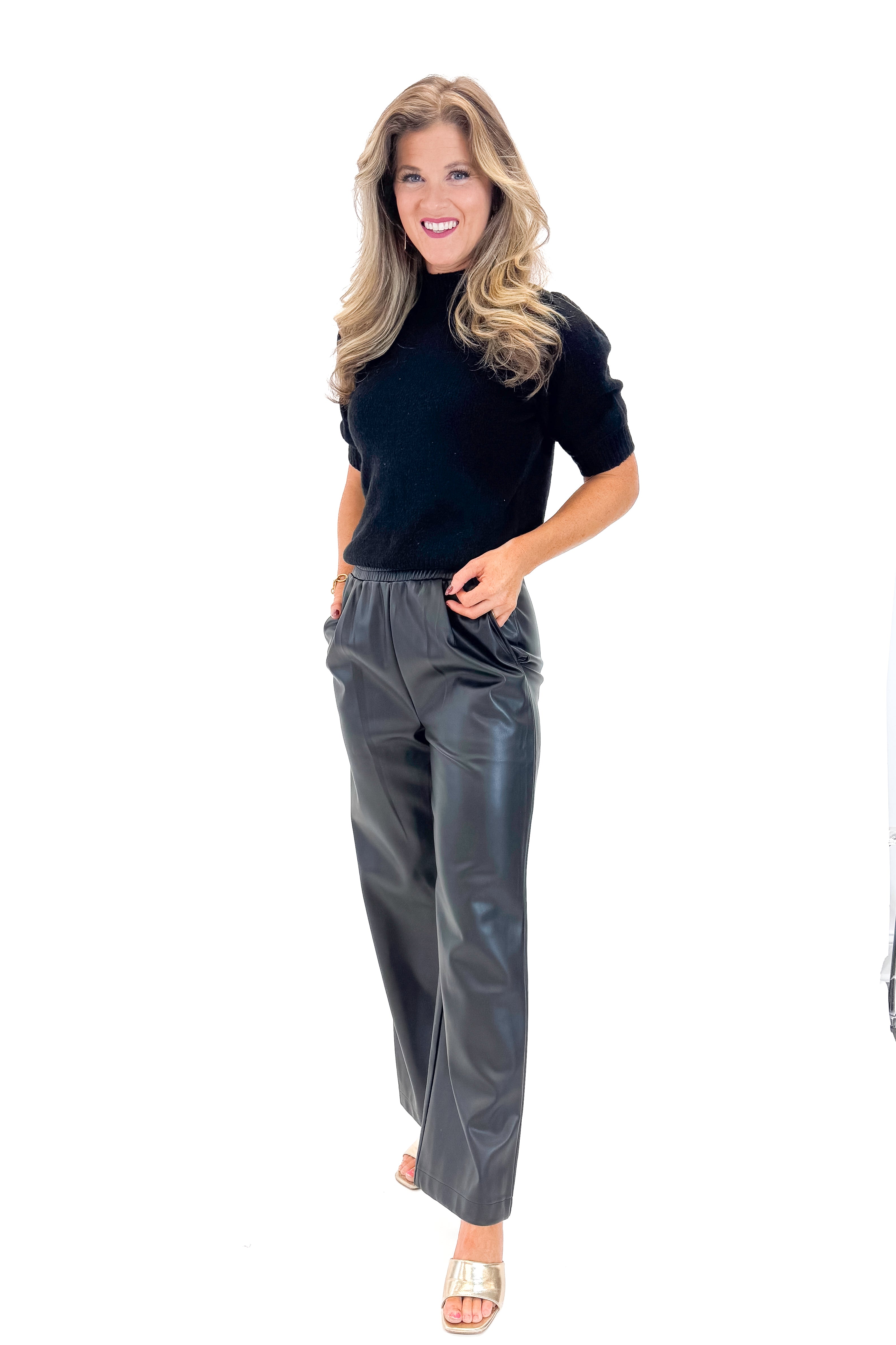 Front view of woman wearing Black Faux Leather Pants with black sweater against white studio background.