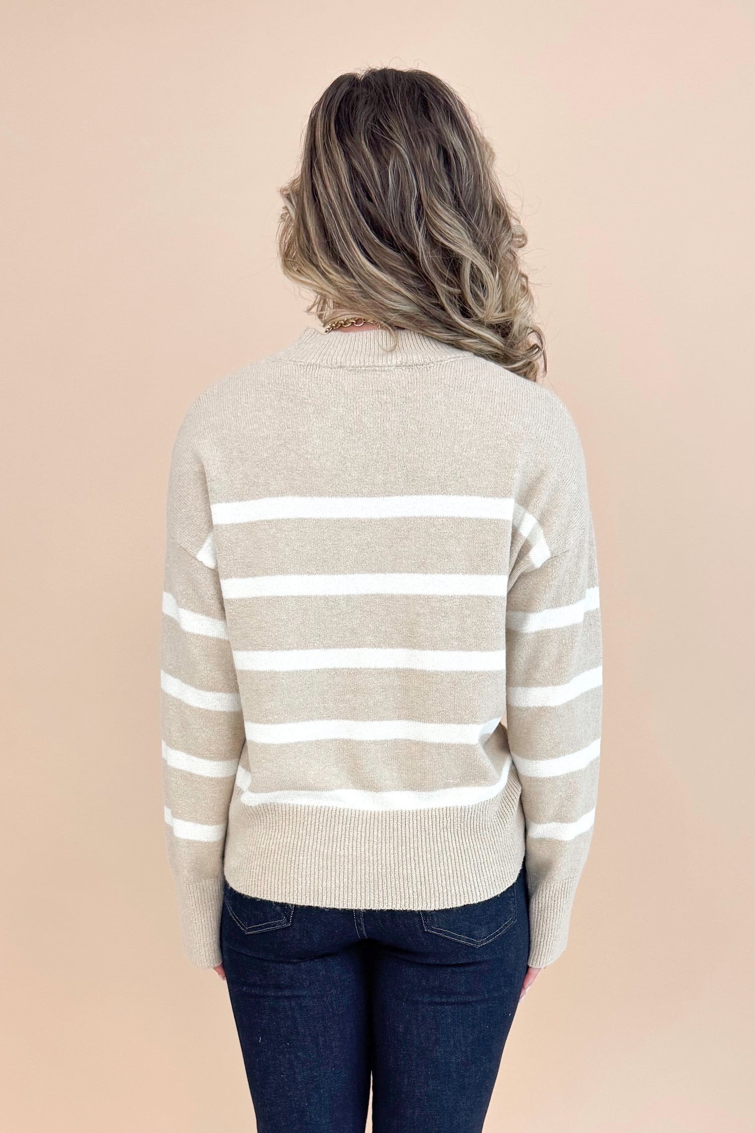 Back view of woman wearing Taupe and Cream Striped Mock Neck Sweater and jeans with tan background.