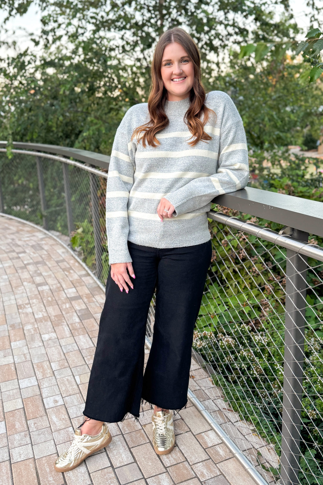 Woman wearing a gray striped sweater and black pants standing on a bridge with greenery in the background